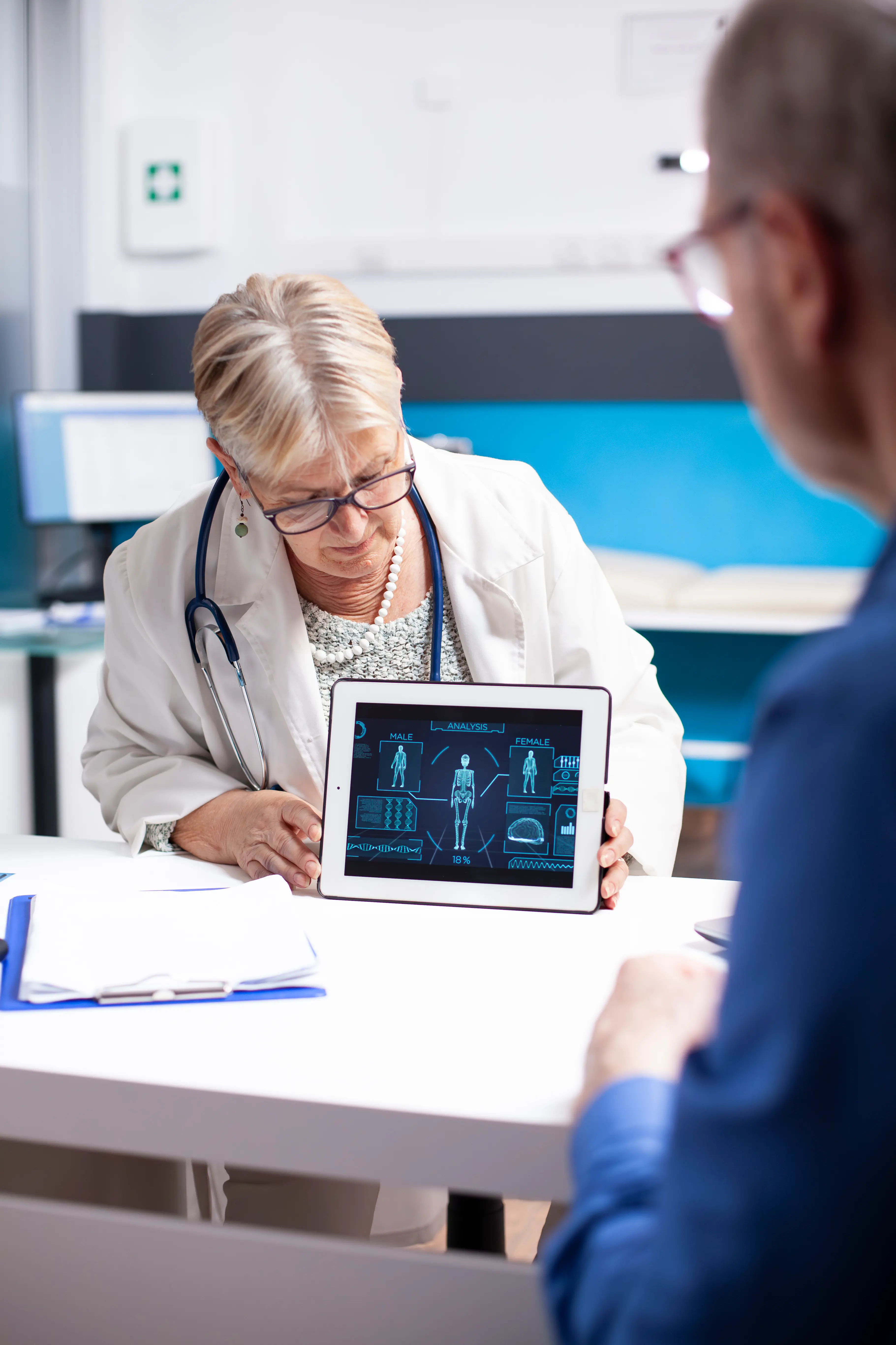 A senior female doctor in a white lab coat and stethoscope showing a digital tablet to a patient. The tablet screen displays a futuristic blue interface with anatomical analysis of a human body, graphs, and medical data. The setting is a bright, professional clinical office