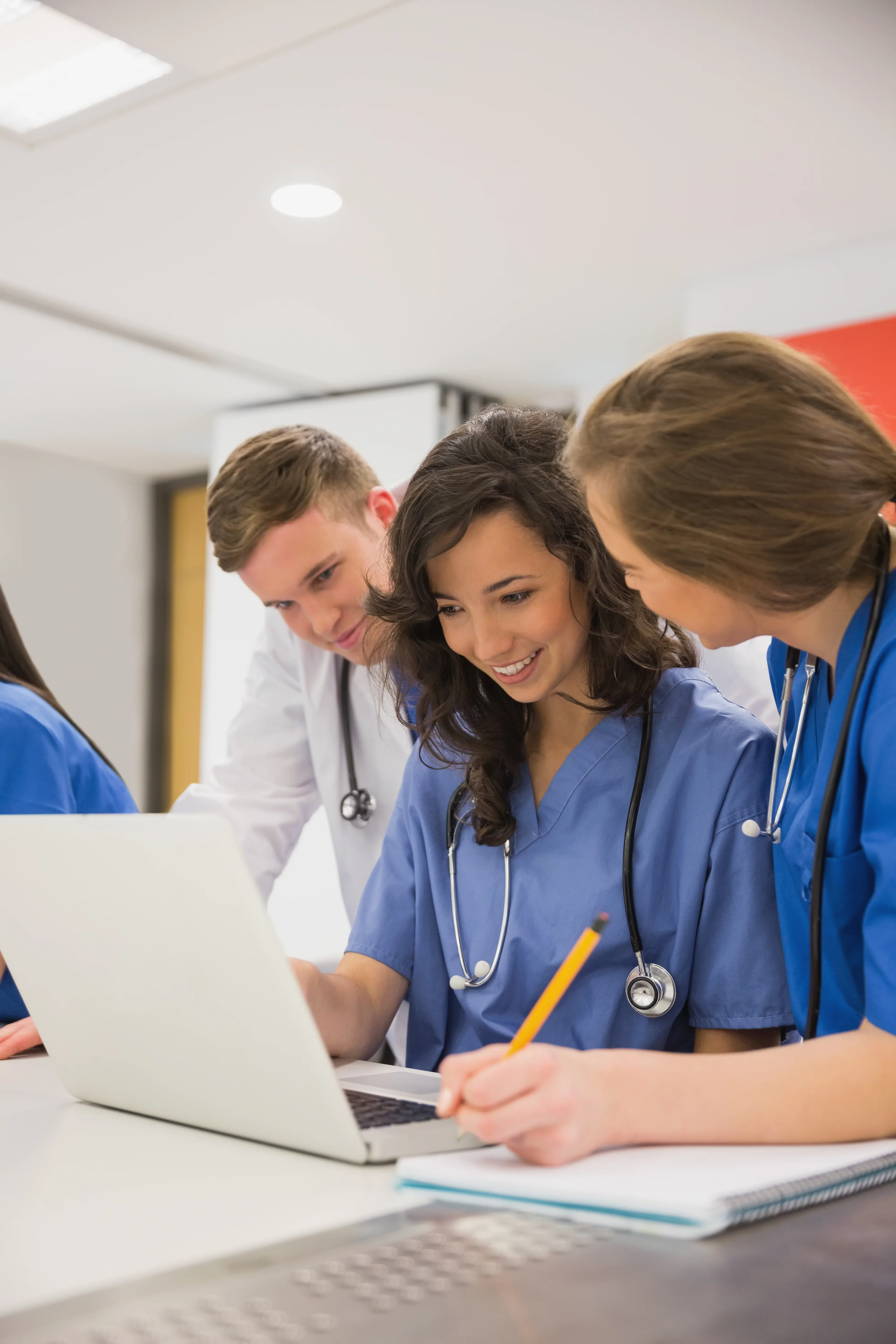 A professional photo of three young medical students or healthcare professionals collaborating in a bright clinical setting. A woman in blue scrubs is seated at a white desk, smiling while looking at a laptop screen. To her right, another woman in blue scrubs leans in to view the screen while taking notes on a notepad with a yellow pencil. Standing behind them, a man in a white lab coat with a stethoscope around his neck looks on. The atmosphere is collaborative and focused on medical education and clinical research.