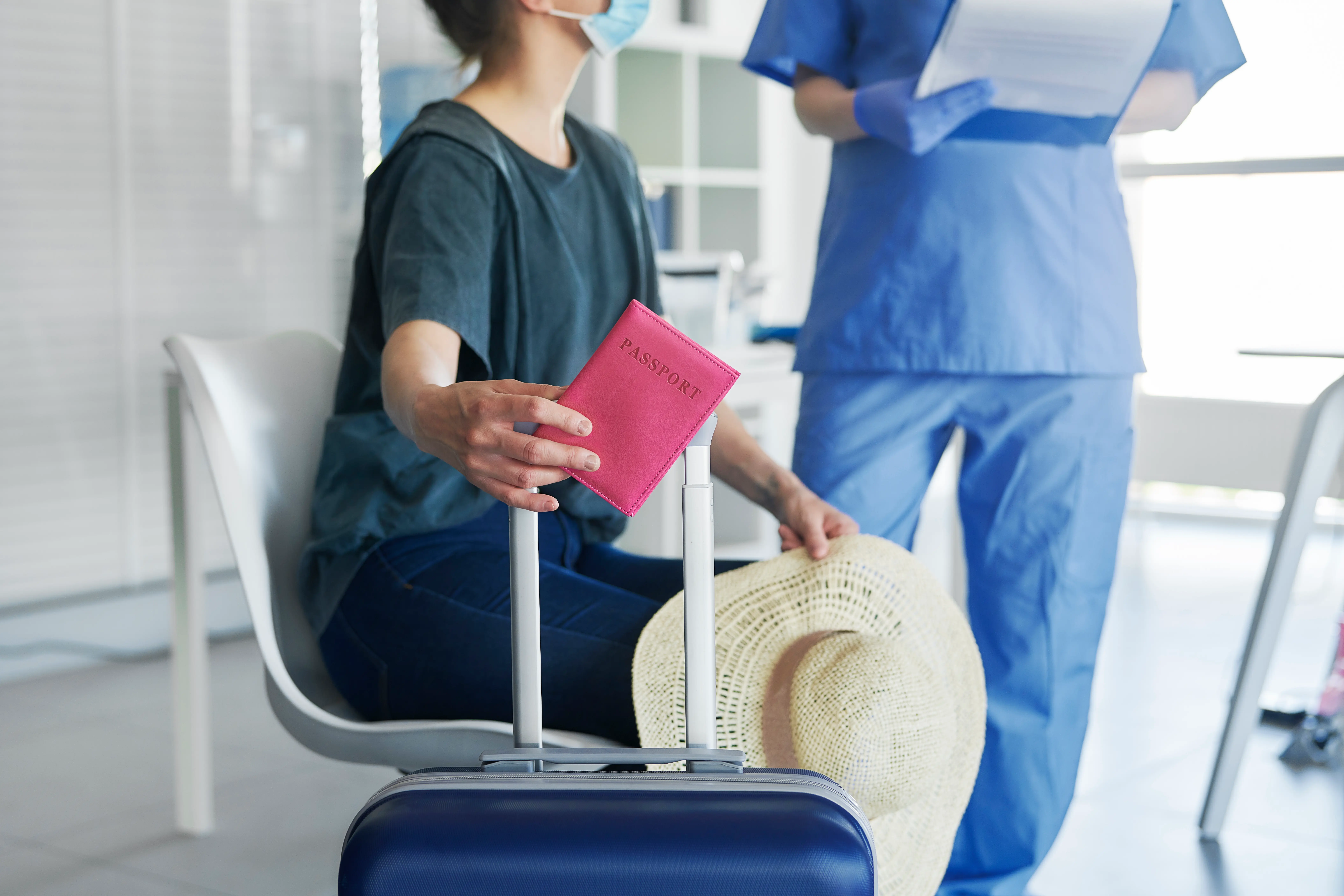 A close-up shot of a person sitting in a waiting area, holding a pink passport cover and a sun hat next to a blue suitcase. In the background, a healthcare professional in blue scrubs and gloves holds a clipboard, suggesting a travel-related medical consultation or document check