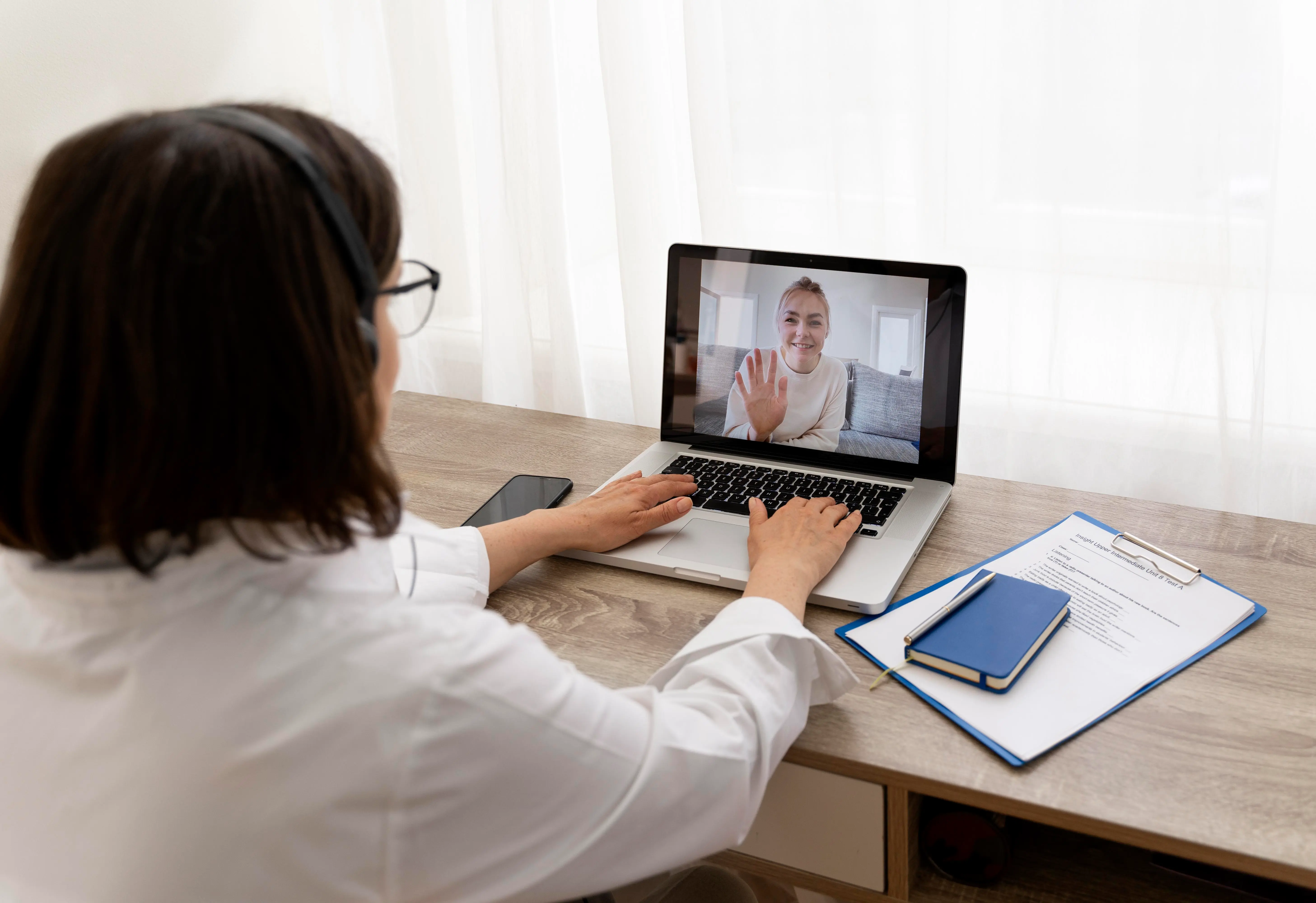 A high-angle, behind-the-shoulder view of a healthcare professional in a white coat wearing a headset, participating in a virtual interview or telehealth session. They are seated at a wooden desk with a laptop that shows a smiling woman waving on the screen. The desk also holds a smartphone, a blue notebook with a pen, and a clipboard with a document